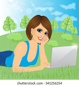 attractive brunette woman lying down on grass using laptop on peaceful park with trees