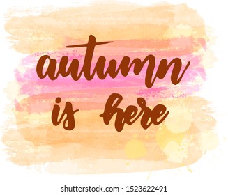 Autumn is here - handwritten modern calligraphy lettering on abstract brushed watercolor background. Season illustration. Orange and pink colored.