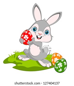 A baby bunny holding a red Easter egg and smiling, more eggs on the ground.
