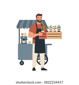A bearded man wearing an apron holds a wooden crate of small plants in front of a mobile coffee cart.