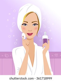 Beautiful blond woman is cleaning her face. Smiling woman in a white bathrobe is caring of her face. Woman is using a cotton pad and a facial skin care product in her bathroom.