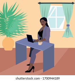 A beautiful girl with dark skin color in a jacket and pants sits at a laptop with a coffee mug. African American in office clothes works at home. Self-isolation during coronavirus quarantine.