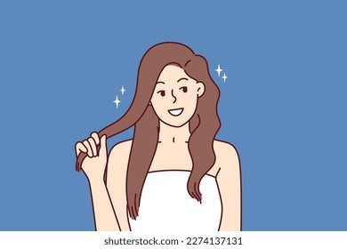 Beautiful girl fixes hair enjoying silky hairstyle after using new herbal shampoo with firming agents. Positive woman in white towel with shiny hair smiling and looking at screen after haircare