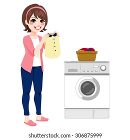 Beautiful happy young brunette woman doing laundry holding clean shirt next to washing machine and basket full of dirty clothes