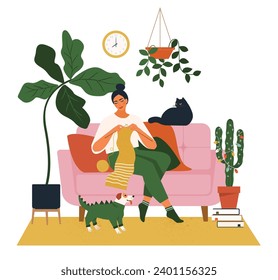 Beautiful woman knitting a scarf on a chair with a cat and a dog. Cozy house with houseplants and cactus with glowing garland.