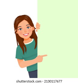 beautiful young woman peeking from behind blank billboard pointing index finger to empty banner for copyspace advertising and announcement messages