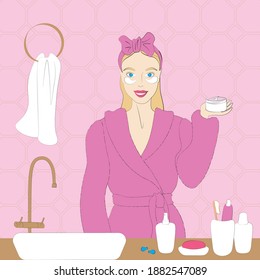 Beauty treatments. A smiling girl in a pink robe is standing in the bathroom. On the hair with a bandage. In the hands of a jar with patches. Next to the sink, cosmetics, bottles, soap.