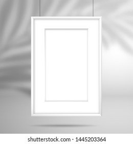 Quadro branco em branco com passagem-partout para uma imagem ou cartaz pendurado em threads. A sombra da folha da palmeira cai na parede atrás. Montagem para design 