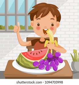 Boy eating fruit. sitting at the table eating Banana. Watermelon and grapes in a tray placed on a table at home in the dining room. emotional mood on child's face feels good. Symbols hand thumb up.