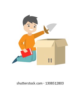 A boy is sitting on the floor and packing his toy sail-boat and a red book in a cardboard box