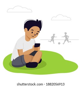 The boy is sitting on the grass in the park. He looks at his phone. His friends are playing football. He has a smartphone addiction, he does not play with them