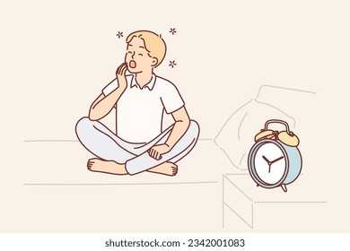 Boy yawns and wants to sleep sitting on bed near alarm clock and feels morning sleepiness due to early rise. Child yawns in need of sleep after hard day at school or long walk with friends.