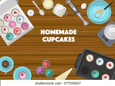 Brown wooden kitchen desk with blue ceramic crockery, kitchen devices, ingredients and coloured cupcakes in the box. Top view.