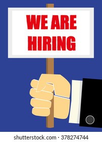 Business man hand in a suit holding a sign or placard with the phrase We Are Hiring in red text