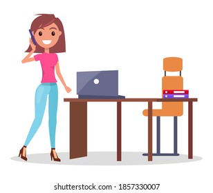 Businesswoman talking by phone standing near office table with laptop and armchair, smiling pretty woman solving business problems, cheerful woman wearing pink t-shirt and jeans using mobile phone