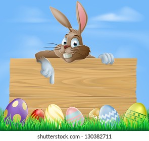 Cartoon Easter bunny pointing at blank wooden sign with painted chocolate Easter eggs in green field