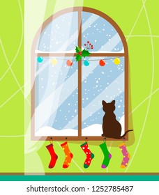 A cat sits on the windowsill by the winter and snow-covered window and looks out the window behind which it is snowing, the window with the curtain is dressed up with a garland and Christmas socks. 