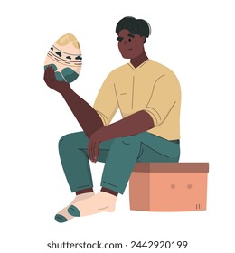The character sits on a box and holds a dyed big Easter egg. The happy man sits in bright clothes and looks at the egg. Modern vector illustration minimal style.