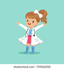 Cheerful baby girl in white coat and stethoscope around her neck standing with hands up isolated on blue background. Child playing doctor game