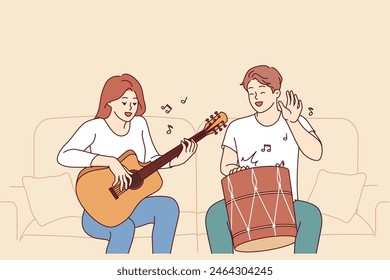 Cheerful friends man and woman playing musical instruments, sitting on sofa and using drum with guitar. Creative guy and girl enjoying playing musical hits during sunday relaxation