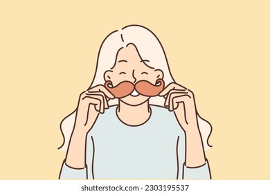 Cheerful girl puts cardboard mustache to face and laughs choosing funny mask for photo. Young woman with fake mustache and playful mood smiling enjoying accessories from photozone at party