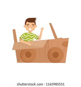 Cheerful little boy sitting in car made of cardboard box and waving hand. Childhood theme. Flat vector design