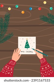 Child's hands with pen writing Christmas card on dark wooden table with garland and pine fir branches. Preparing for winter holidays background. Top view flat design illustration