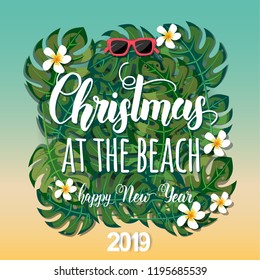 Christmas background with palm leaves, sunglasses, tropical flowers and hand made trendy lettering. Hand written quote "Christmas at the beach. Happy New Year 2019"