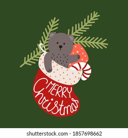 Christmas card. A gift sock with a teddy bear, candy and a gift hanging on a spruce branch. Lettering MERRY CHRISTMAS.