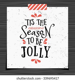 Christmas typographic design greeting card template. 'Tis the Season to be Jolly message in black and red on white background. Christmas card with a strip of washi tape on dark wood background.