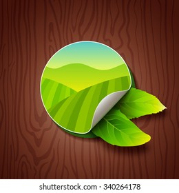 Circle Label with Green Fields, Mountains and Two Tea Leaves on Wood Background. Tea Garden on Hills Illustration. Colorful Sticker for Packaging, Logo and Web Design.