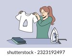 Clumsy housewife ironing clothes burnt hole in t-shirt and dont know how to fix mistake. Woman stands near ironing board with clothes and iron and looks at damaged item in confusion.