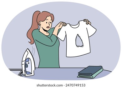 Clumsy housewife ironing clothes burnt hole in t-shirt and dont know how to fix mistake. Woman stands near ironing board with clothes and iron and looks at damaged item in confusion.