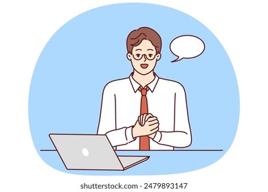 Confident businessman is sitting at table with laptop and looking at camera recording business training. Guy in business clothes with dialogue cloud works as teacher online training courses