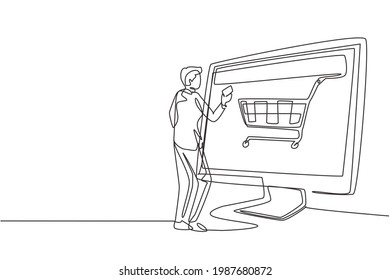 Continuous one line drawing young man shopping online via giant computer screen with shopping cart inside. Digital lifestyle, consumerism concept. Single line draw design vector graphic illustration