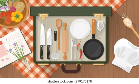 Cooking and kitchen utensils in an open vintage suitcase on a checked tablecloth with chef's hat, cookbook and fresh vegetables