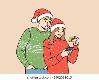 Couple in christmas clothes makes purchases on phone mobile app and uses credit card to pay for goods online. Christmas man and woman holding friend business card and dialing number in smartphone