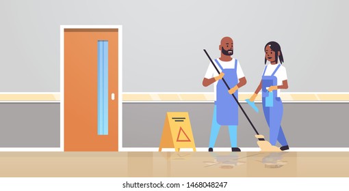 couple cleaners in uniform working together cleaning service concept african american man woman janitors using mop spray plastic bottle modern clinic corridor interior full length flat