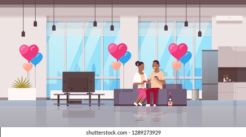 couple drinking wine happy valentines day concept african american man woman lovers watching tv on couch modern apartment home interior air heart shape balloons flat horizontal