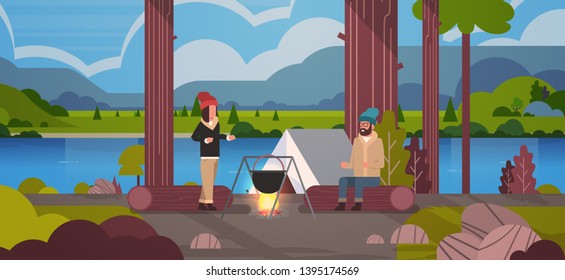 couple hikers sitting on log man woman cooking meals in bowler boiling pot at campfire near camp tent camping concept landscape nature river mountains background horizontal full length