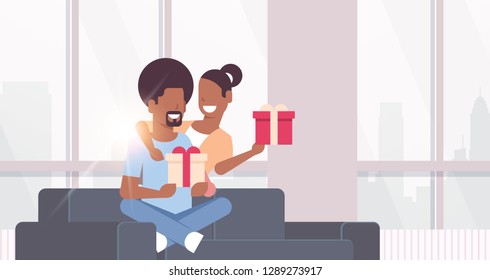 couple holding present wrapped gift boxes african american man woman embracing sitting couch celebrating holiday concept happy lovers modern living room interior horizontal