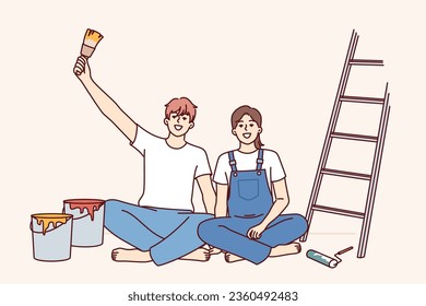 Couple makes repairs in apartment with own hands and sits on floor near ladder and buckets of paint for painting walls. Cheerful man and woman rejoice at opportunity to make diy repairs in room