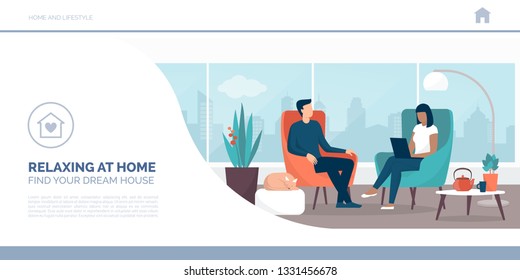 Couple relaxing at home sitting on the armchairs  in the living room, the woman is connecting with her laptop, lifestyle and interior design concept