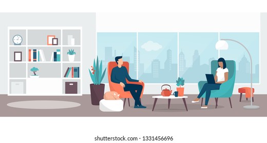 Couple relaxing at home sitting on the armchairs  in the living room, the woman is connecting with her laptop, lifestyle and interior design concept