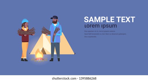 couple tourists hikers holding firewood african american man woman organizing fire camp with canvas tent and campfire hiking camping concept horizontal full length copy space flat