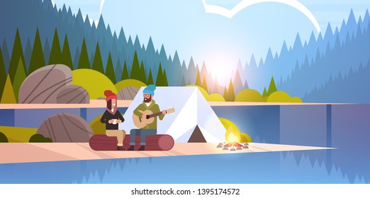 couple tourists hikers relaxing in camp man playing guitar for girlfriend sitting on log hiking concept sunrise landscape nature river forest mountains background flat horizontal full length