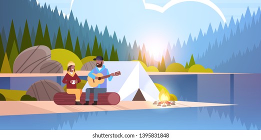 couple tourists hikers relaxing in camp african american man playing guitar for girlfriend sitting on log hiking concept sunrise landscape nature river forest mountains background horizontal