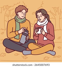 Couple in warm clothes sits on ground among fallen leaves, pouring hot tea from thermos. Peaceful autumn moment with cozy atmosphere