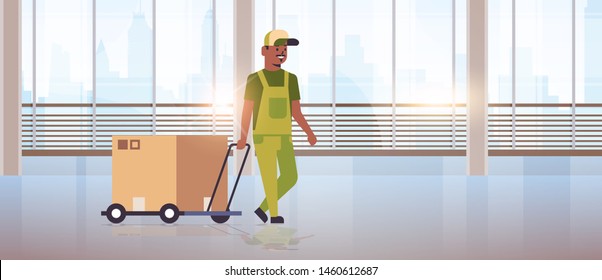 courier in uniform pushing trolley with cardboard box express delivery service concept african american worker with hand truck modern office hall interior flat full length horizontal