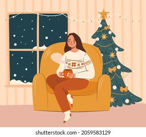 Cozy Christmas evening concept. Girl in ugly sweater sitting in armchair with hot drink. Snowy weather, christmas tree in the background